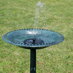 A round black metal bird bath fountain with water spraying upward, set on a green grass lawn.